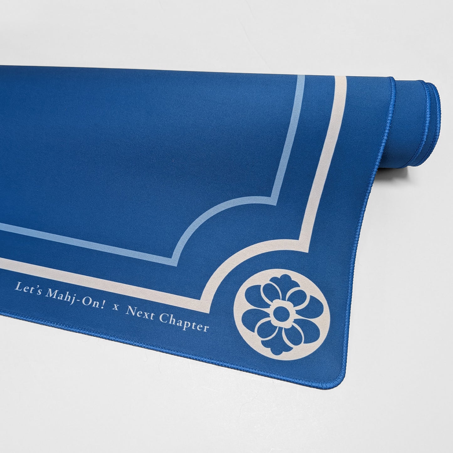 A blue Mahjong playmat with white printed decorative lines, a floral emblem, and the text 'Let's Mahj-On! x Next Chapter', shown partially unrolled on a light gray background.