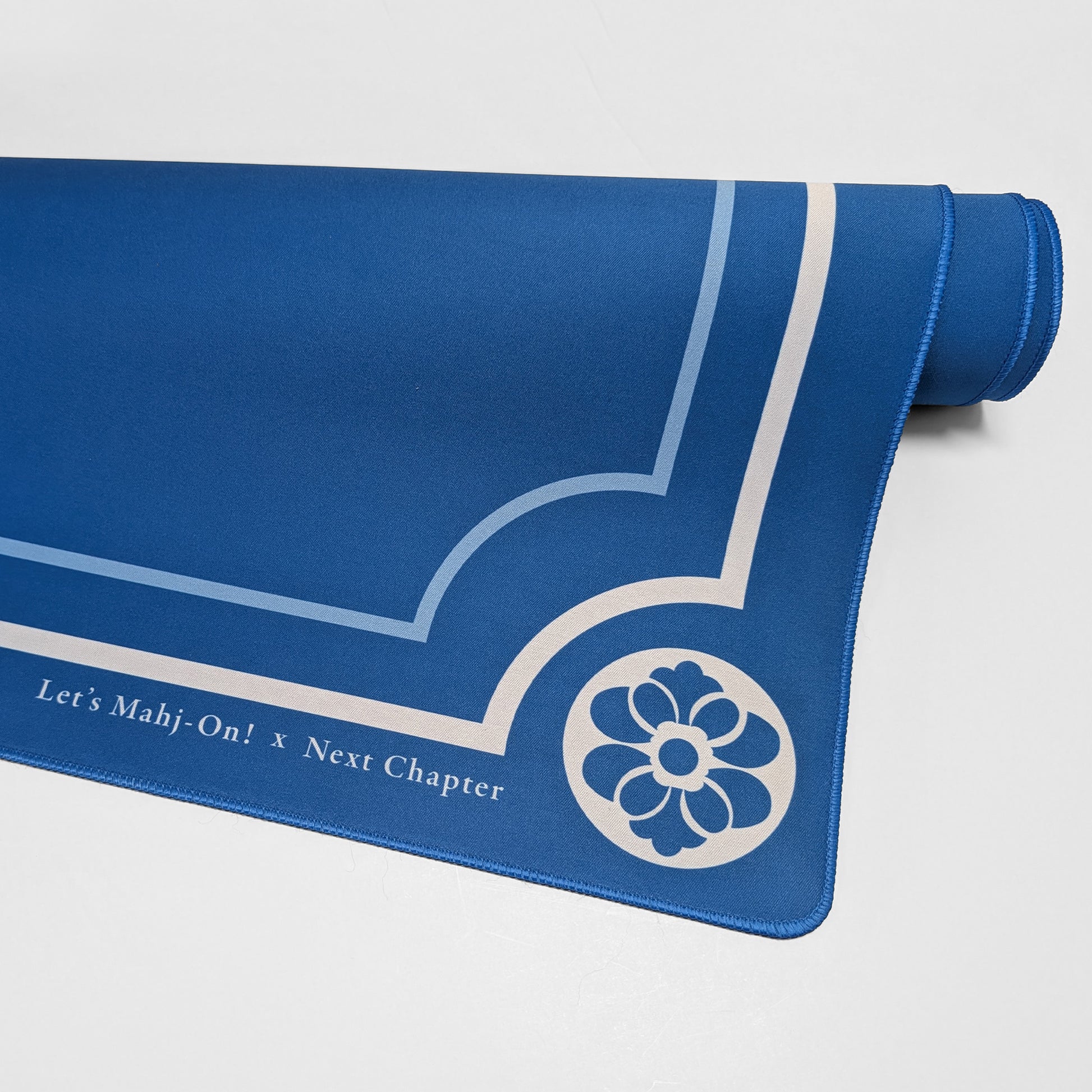 A blue Mahjong playmat with white printed decorative lines, a floral emblem, and the text 'Let's Mahj-On! x Next Chapter', shown partially unrolled on a light gray background.