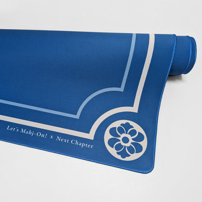 A blue Mahjong playmat with white printed decorative lines, a floral emblem, and the text 'Let's Mahj-On! x Next Chapter', shown partially unrolled on a light gray background.