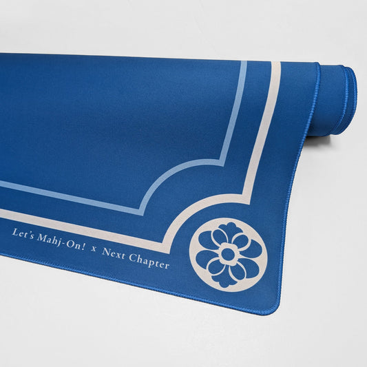 A blue Mahjong playmat with white printed decorative lines, a floral emblem, and the text 'Let's Mahj-On! x Next Chapter', shown partially unrolled on a light gray background.