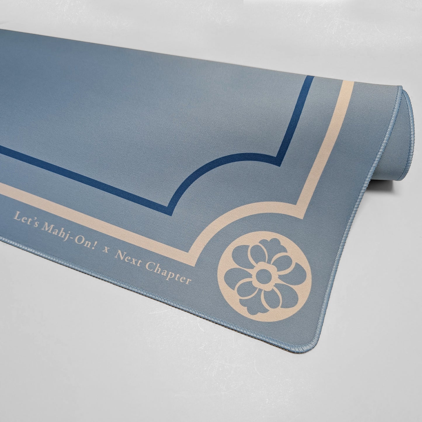 A rolled-up Mahjong playmat in a dusty slate blue color, featuring navy blue geometric line art and a beige floral emblem. Text along the bottom edge reads 'Let's Mah-j-On! x Next Chapter'.