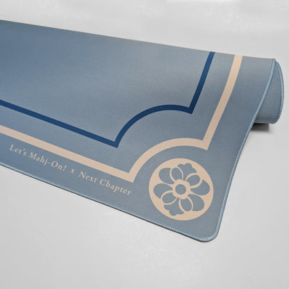 A rolled-up Mahjong playmat in a dusty slate blue color, featuring navy blue geometric line art and a beige floral emblem. Text along the bottom edge reads 'Let's Mah-j-On! x Next Chapter'.