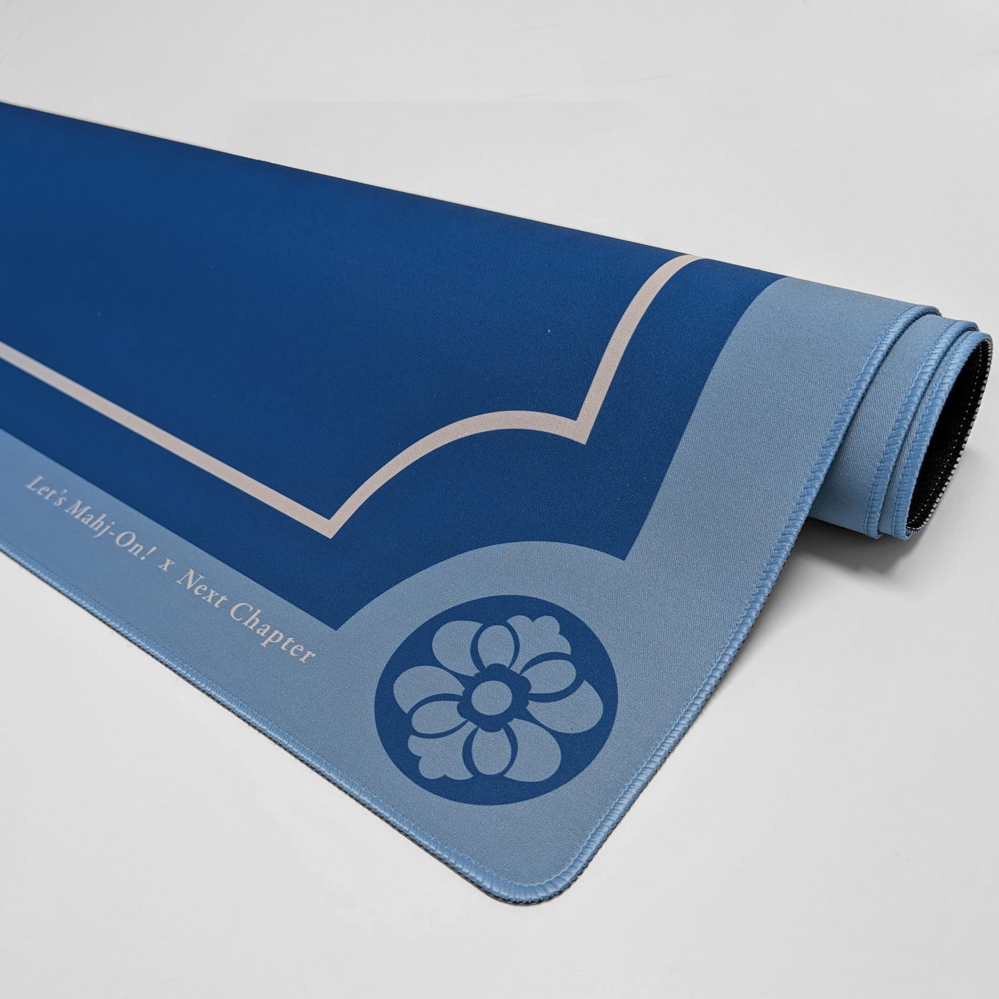 A rolled-up Mahjong playmat in shades of blue, featuring a light blue floral emblem and the text 'Let's Mahj On! X Next Chapter' printed on the side, photographed on a neutral light gray background.