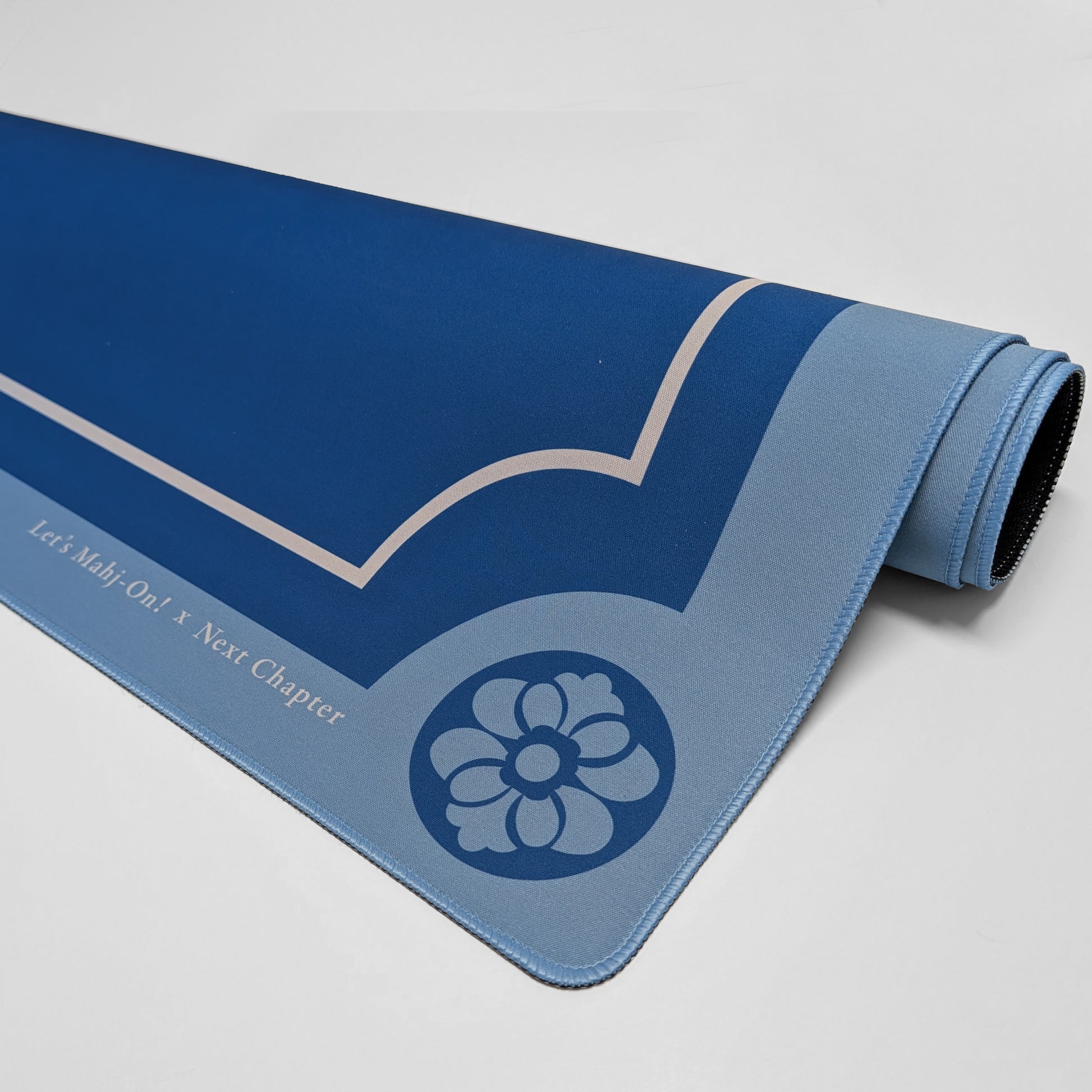A rolled-up Mahjong playmat in shades of blue, featuring a light blue floral emblem and the text 'Let's Mahj On! X Next Chapter' printed on the side, photographed on a neutral light gray background.